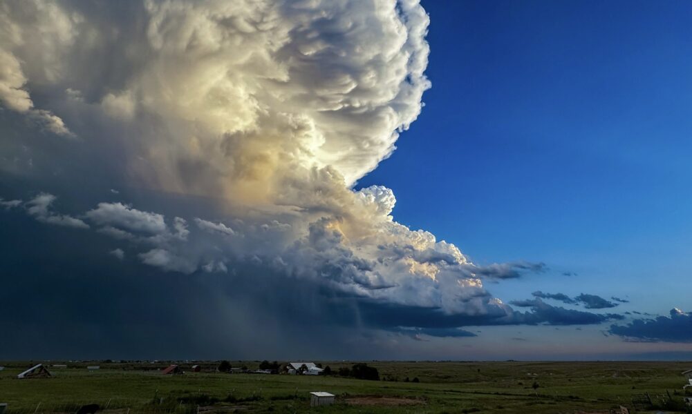 Dramatic cloud formation with vibrant blue sky and hints of sunlight peeking through, set against a rural landscape with fields and scattered buildings. Ideal for weather enthusiasts and landscape photography.