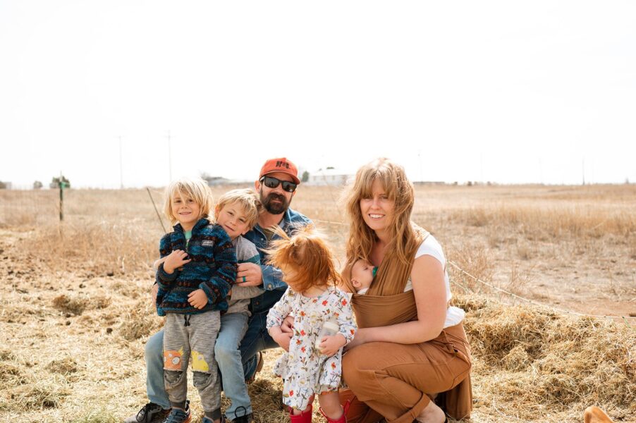 Family portrait outdoors in a rural setting, featuring a mother holding a baby in a wrap, surrounded by three children and a father. The background shows a dry landscape with sparse vegetation and power lines. The family is dressed casually, enjoying a sunny day together.